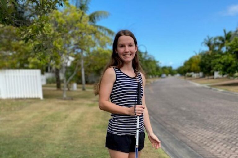 A young adult standing outside with their white cane. They are looking at the camera smiling.