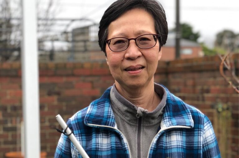 A person standing outside smiling at the camera while holding their white cane.