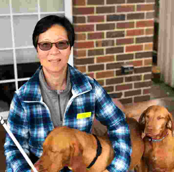 A person seated next to their two dogs. They are also holding their white cane and are smiling at the camera.