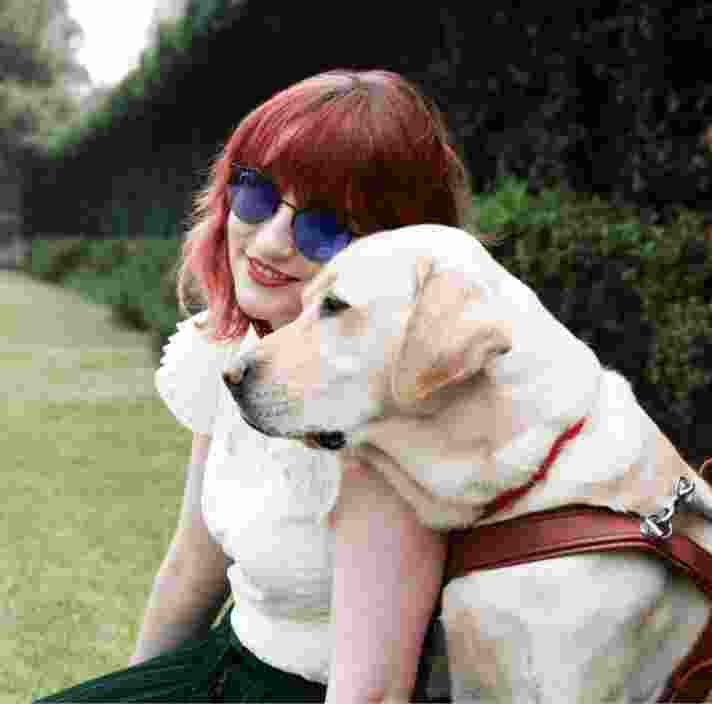 A person and a yellow Guide Dog, in harness, sitting on grass outside. The person is smiling at the camera and the Guide Dog is a profile view.