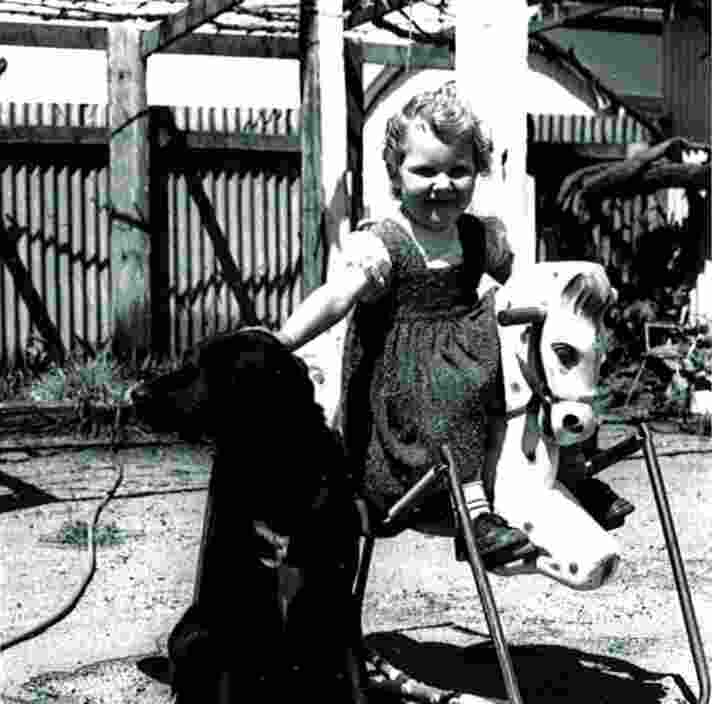 1940's Black and white photograph of a child riding a toy horse whilst patting a black labrador dog that is next next to them. The child has a smile on their face.