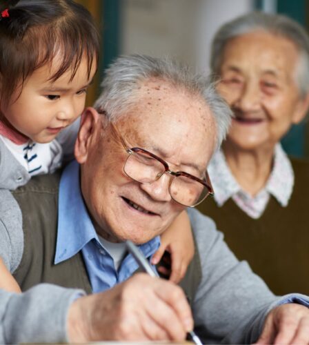 An older man writing while a young girl puts her arms around his shoulders, hugging him. The man is smiling and there is an older woman in the background who is not in focus but is also smiling.