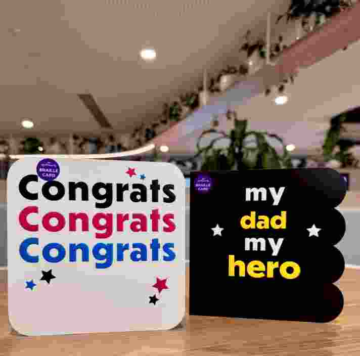 Two Hallmark Celebration cards with braille on the outside, sit standing up on a wooden table facing the camera. One reads 'Congratulations' and one reads 'My dad, my hero.'