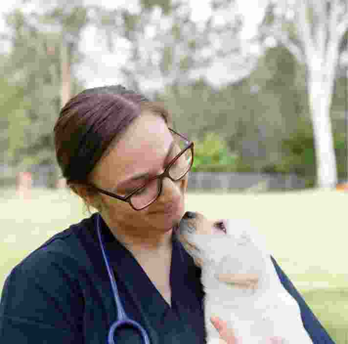 A veterinarian holding an eight week old yellow labrador puppy. The photo is taken outside and the puppy and vet are looking at each other, the veterinarian is smiling at the puppy.