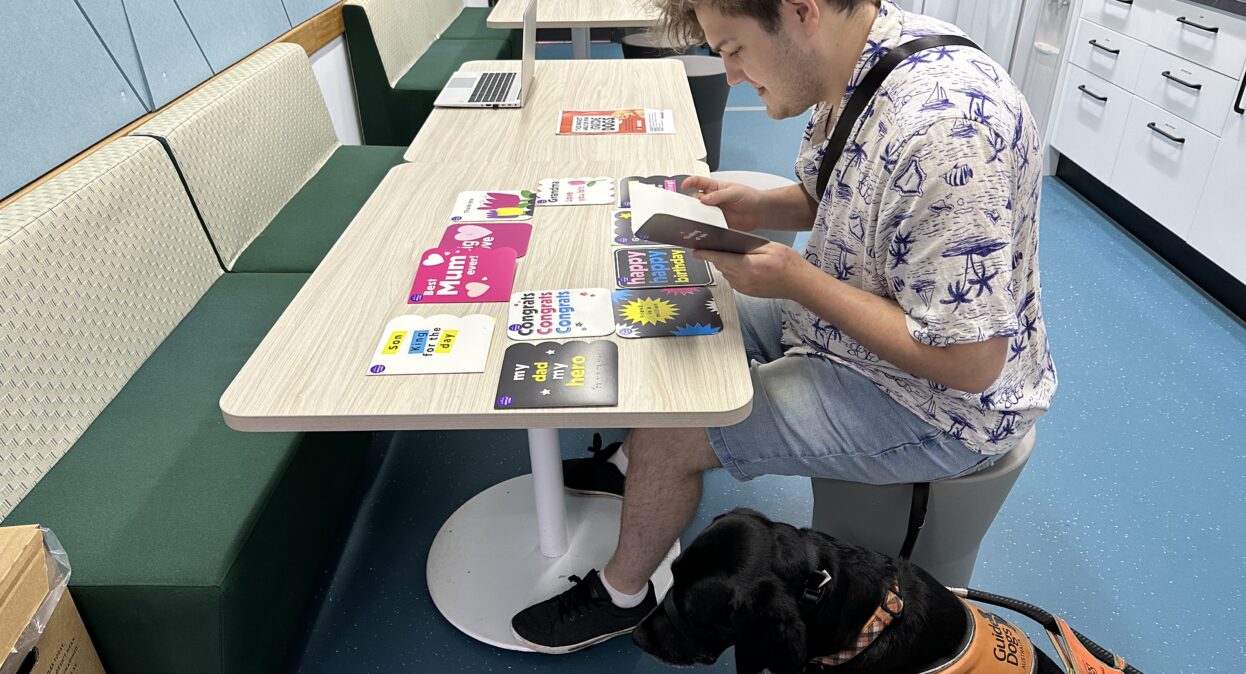 A male reads a braille birthday card sitting at a table. His Guide Dog sits beside him on the floor.