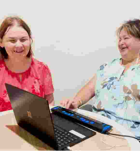 Two people laughing as they work together at a computer with a braille reader