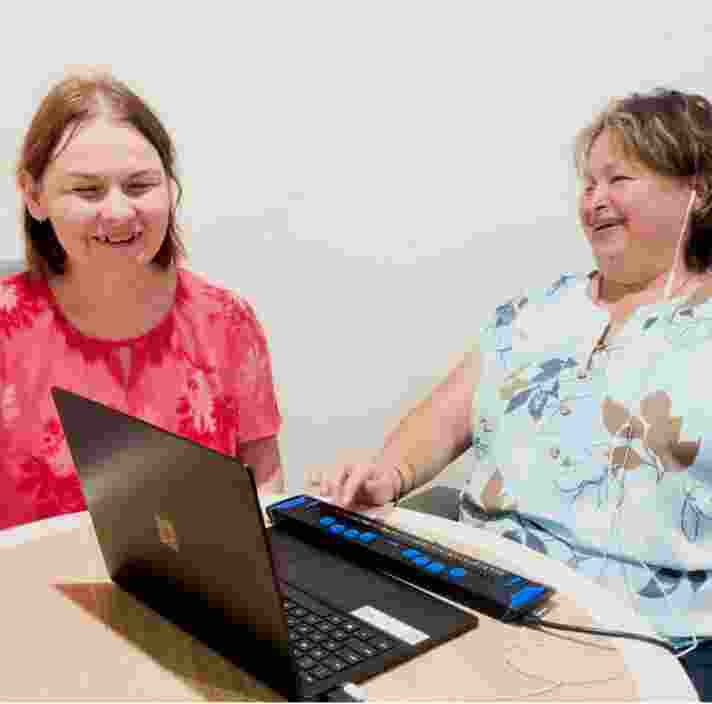 Two people laughing as they work together at a computer with a braille reader