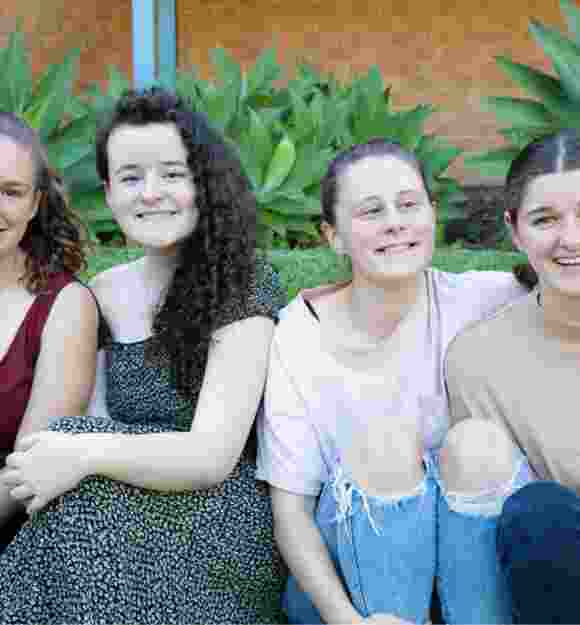 A group of four young adults sitting outside on the grass all in a row. Everyone is looking at the camera smiling.