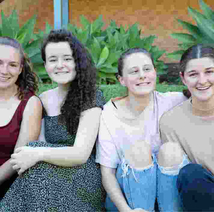 A group of four young adults sitting outside on the grass all in a row. Everyone is looking at the camera smiling.
