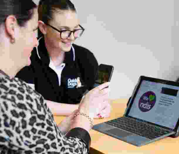 A person and a Guide Dog's staff member looking at a laptop. The person is holding up an Iphone to the laptop and the staff member is smiling.