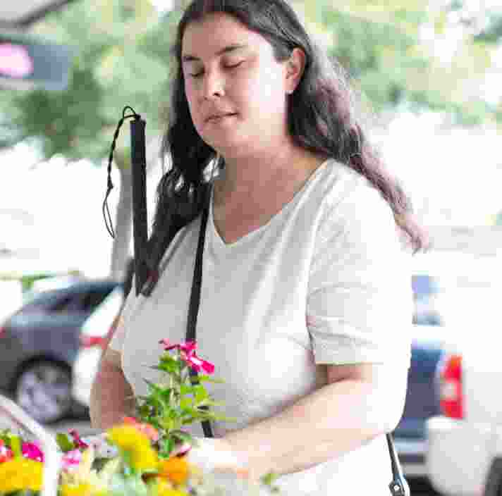 A person is standing in the street holding some flowers in one hand and their white cane in their other arm.
