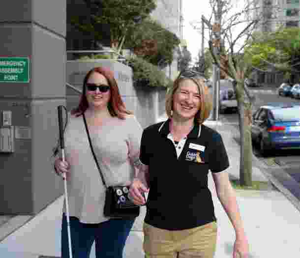 A person with their white cane walking down the street with a Guide Dogs staff member. Both people are smiling at the camera and the person with the cane is holding the staff member by the elbow.