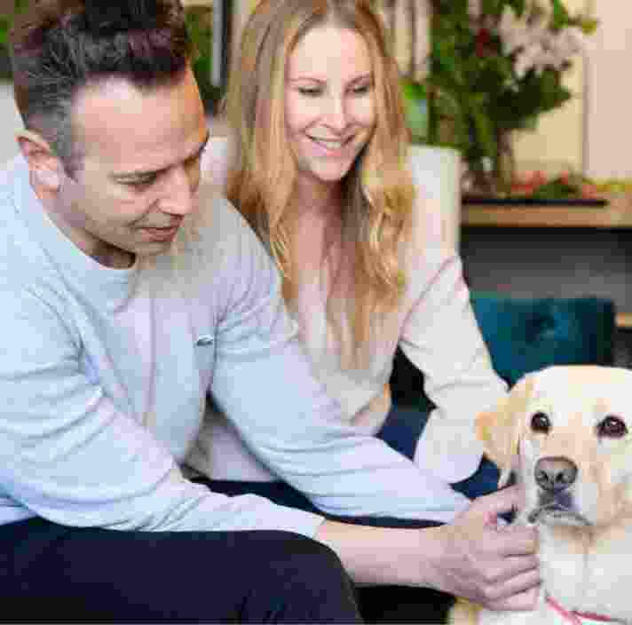 Two people sitting on their couch patting their yellow labrador dog.