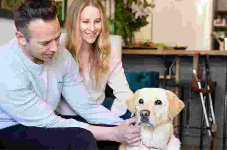 Two people sitting on their couch patting their yellow labrador dog.