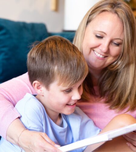 Young child sitting on the ground with their mother next to them. They are both smiling and looking at an Ipad.