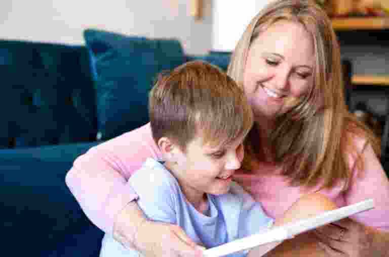 Young child sitting on the ground with their mother next to them. They are both smiling and looking at an Ipad.