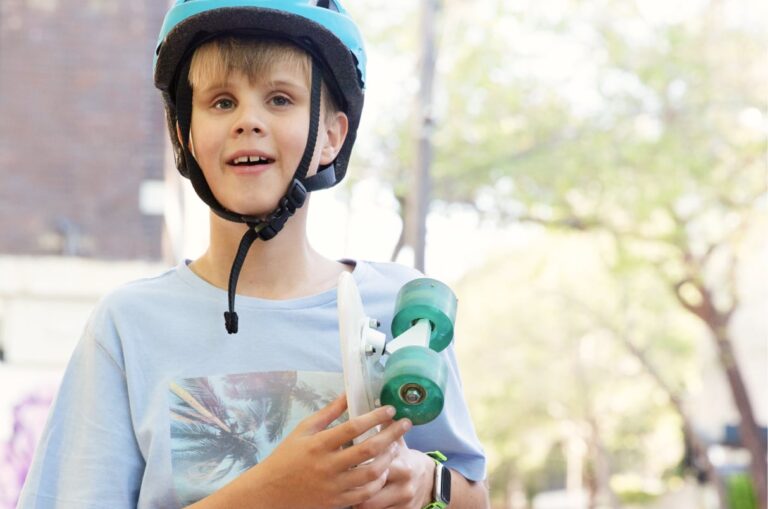 Young child wearing a helmet, holding a skateboard and standing outside in the city streets. The child is looking at the camera and smiling.