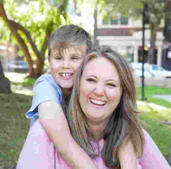 Young child with their arms around their mothers neck and they are both looking at the camera smiling. They are seated on grass outside.