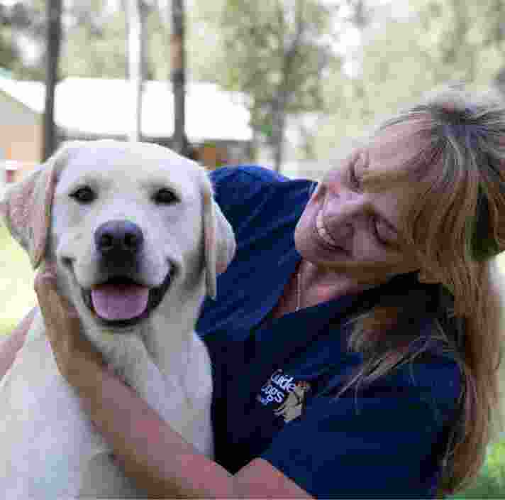 A veterinarian and fully grown yellow labrador are sitting outside. The veterinarian is looking at the dog with a smile on their face and the dog is smiling at the camera.