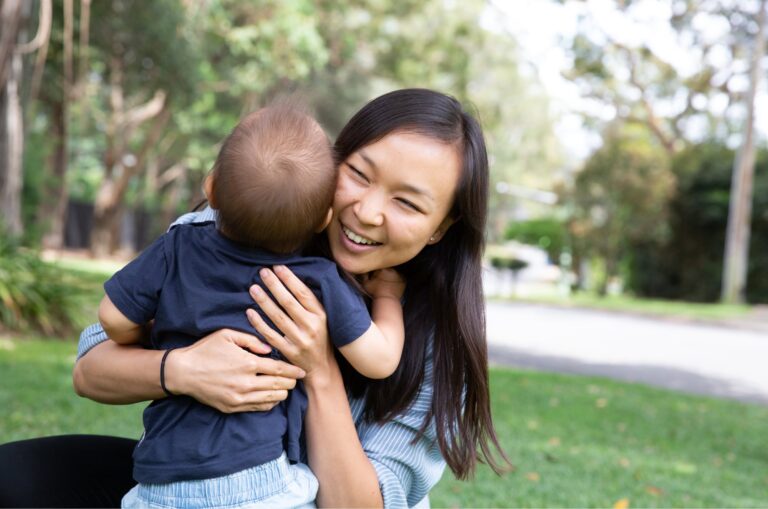 A mother and baby outside on the grass hugging each other. The mother is smiling and the baby has its back to the camera.