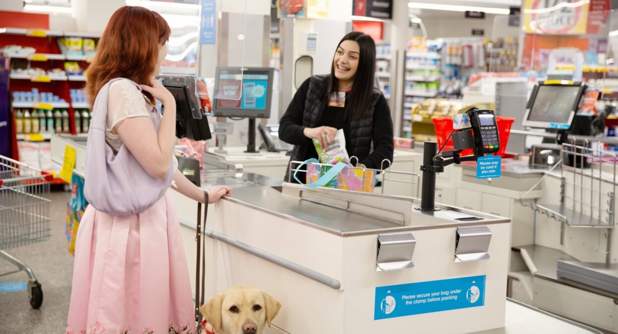 A person at a check out who is looking at the cashier. The cashier is smiling. The person at the checkout has a yellow labrador Guide Dog in harness who is looking at the camera.