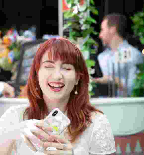 An adult sitting outside at a cafe laughing while holding their mobile phone.