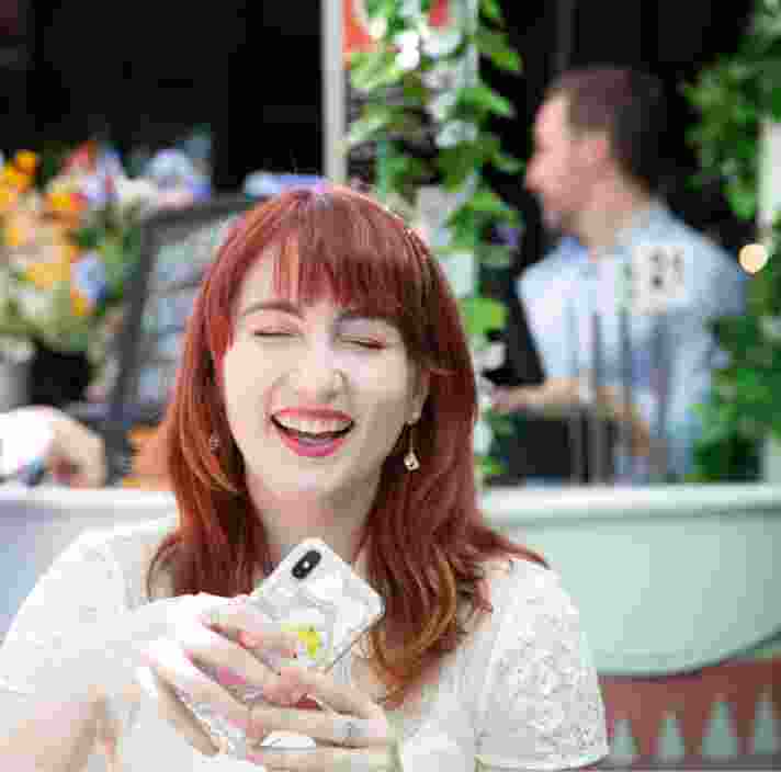 An adult sitting outside at a cafe laughing while holding their mobile phone.