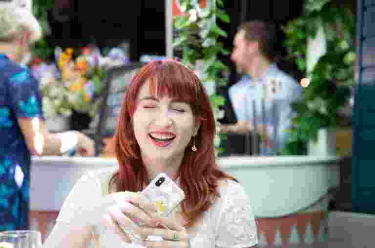 An adult sitting outside at a cafe laughing while holding their mobile phone.
