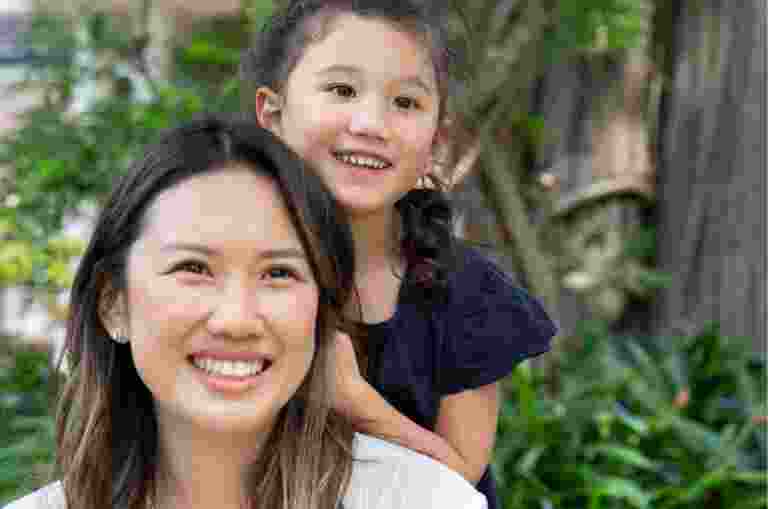 A mother and young child outside smiling at the camera. The young child is standing behind the mother with their hands on her back.