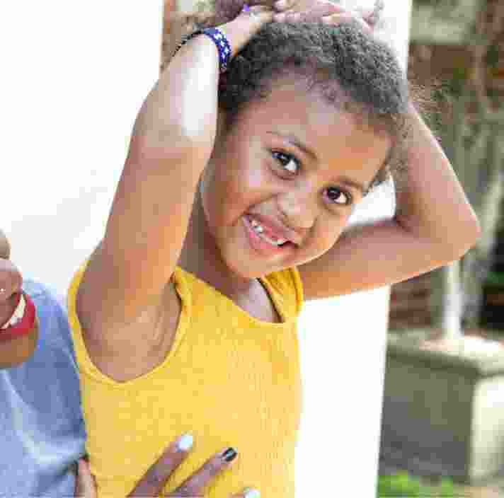 A young child smiling at the camera while their mother looks at them from behind. The mother is smiling at their child.