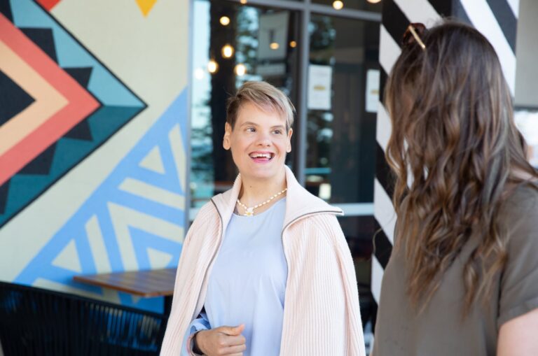 Two people looking at each other while they are walking down the street. One has their back to the camera while the other is smiling