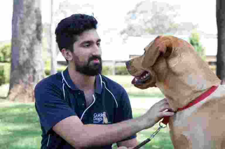 A Guide Dogs staff member and a caramel labrador dog sitting outside. The staff member and dog are both looking at each other.