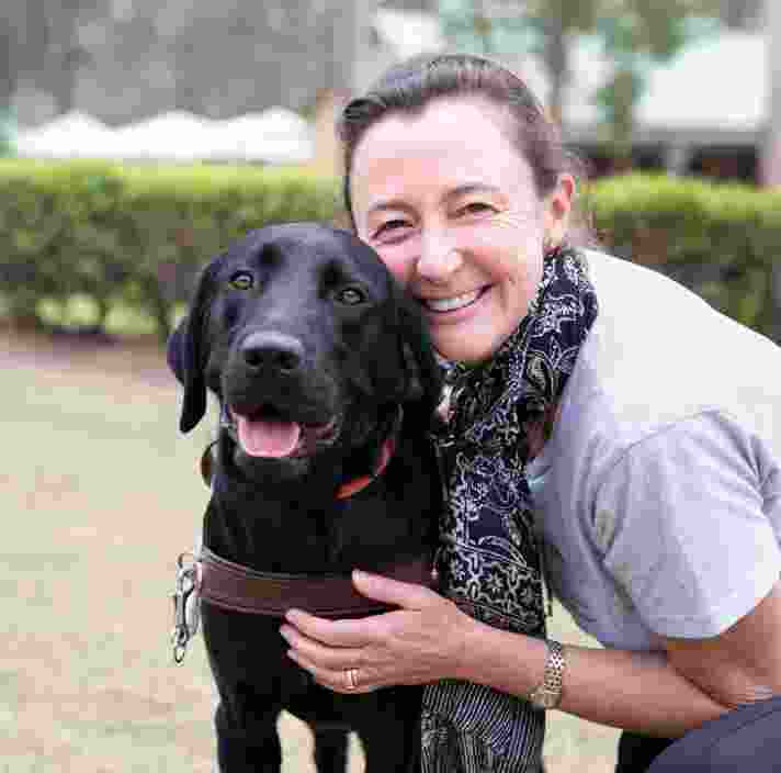 A person hugging a black labrador Guide Dog in harness. The person and dog are looking at the camera and the person is smiling,