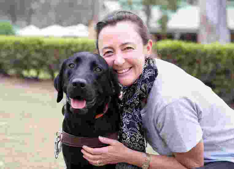 A person hugging a black labrador Guide Dog in harness. The person and dog are looking at the camera and the person is smiling,