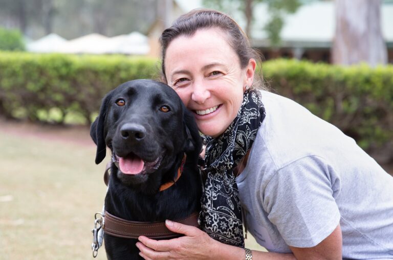 A person hugging a black labrador Guide Dog in harness. The person and dog are looking at the camera and the person is smiling,