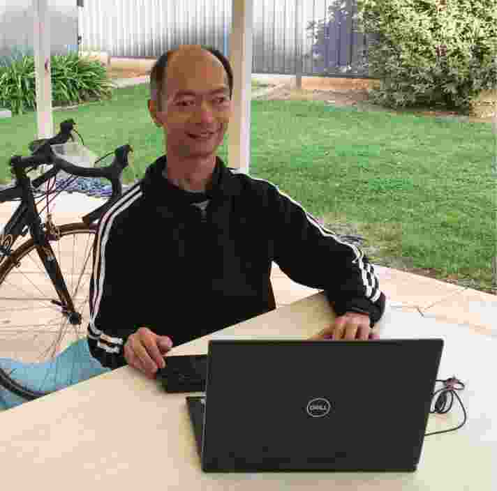 A person sitting outside at a table with their laptop. The person is smiling at the camera.