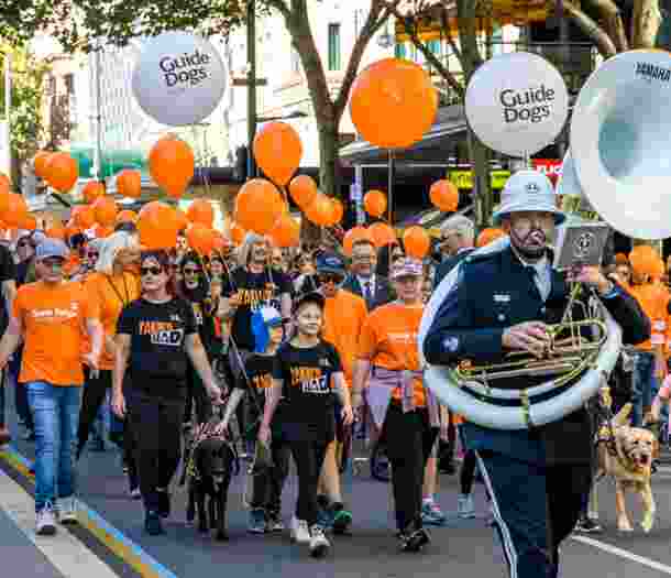 A large number of Guide Dogs staff and supporters walking in a parade. They are all wearing branded Guide Dogs orange t-shirts and some have orange branded helium balloons. At the front of the parade is a person playing a large french horn.