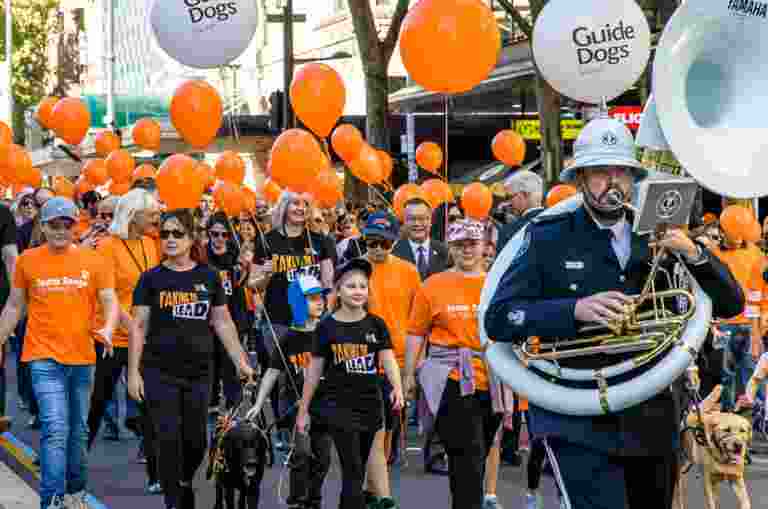 A large number of Guide Dogs staff and supporters walking in a parade. They are all wearing branded Guide Dogs orange t-shirts and some have orange branded helium balloons. At the front of the parade is a person playing a large french horn.