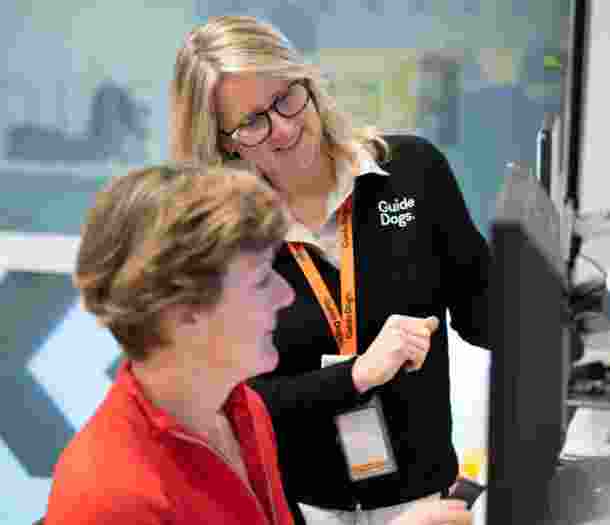 A person is sitting at a computer with a Guide Dogs staff member standing beside them. The Guide Dogs staff member is showing the person sitting at the computer how to it.