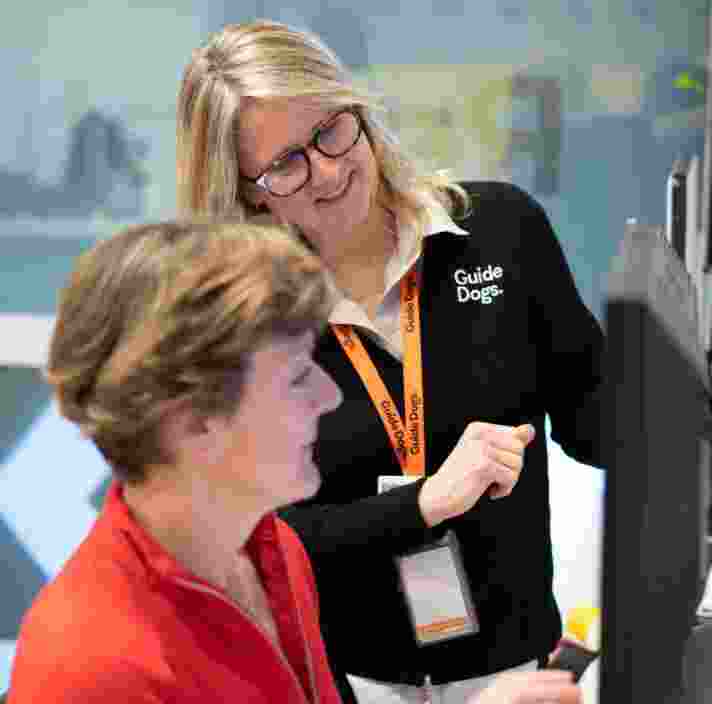 A person is sitting at a computer with a Guide Dogs staff member standing beside them. The Guide Dogs staff member is showing the person sitting at the computer how to it.