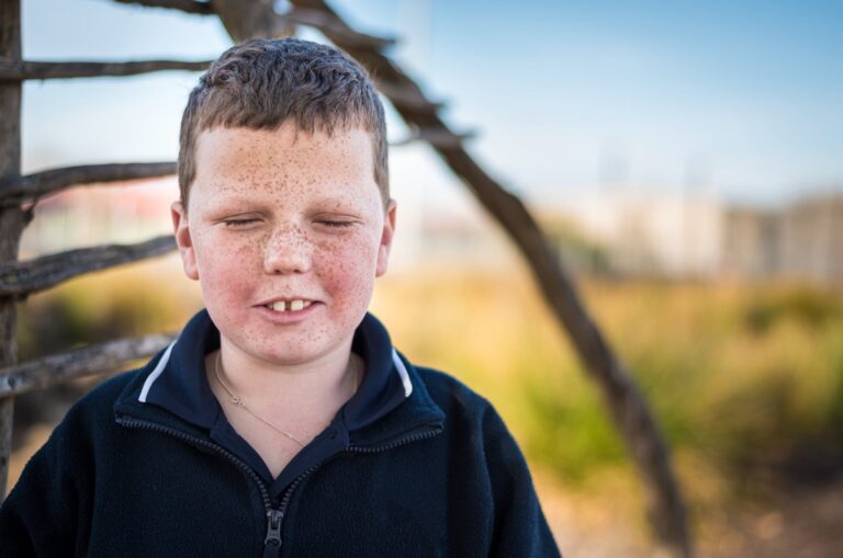 A young child standing outside looking at the camera. The child has their eyes closed.
