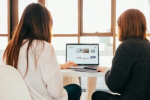 Two women using a laptop computer, one has her hand on the trackpad