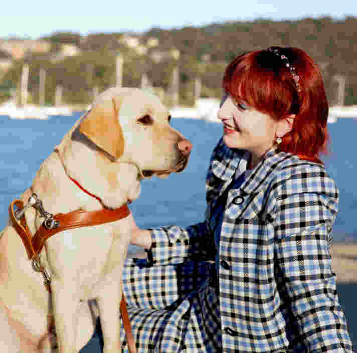 A person sitting by the water smiling at their yellow Labrador guide dog