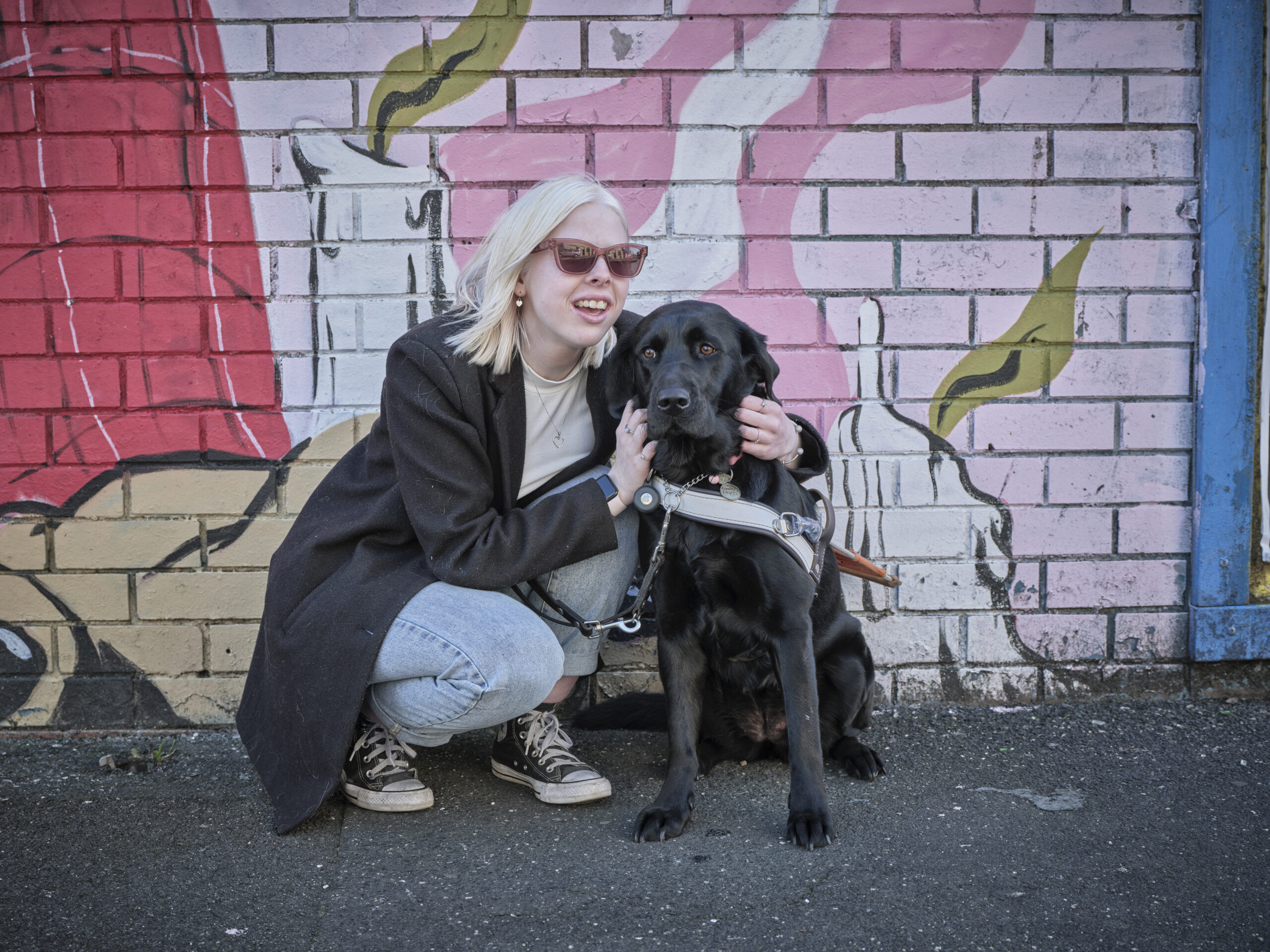 A Guide Dog Client adn her dog crouch down in front of a a colourful wall and have a cuddle