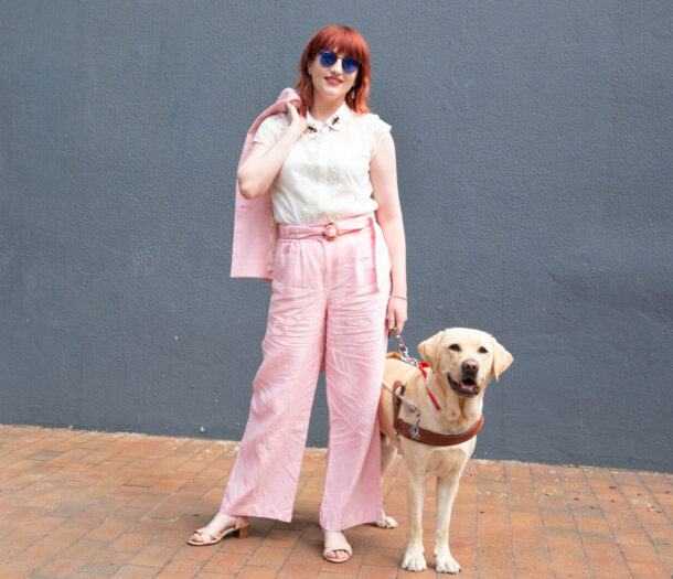 A woman stands elegantly posed in a pink suit with her bright eyed Guide Dog.