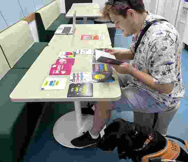 A male with low vision or blindness reads a braille card while sitting down at a table. Their Guide Dog sits at their feet.