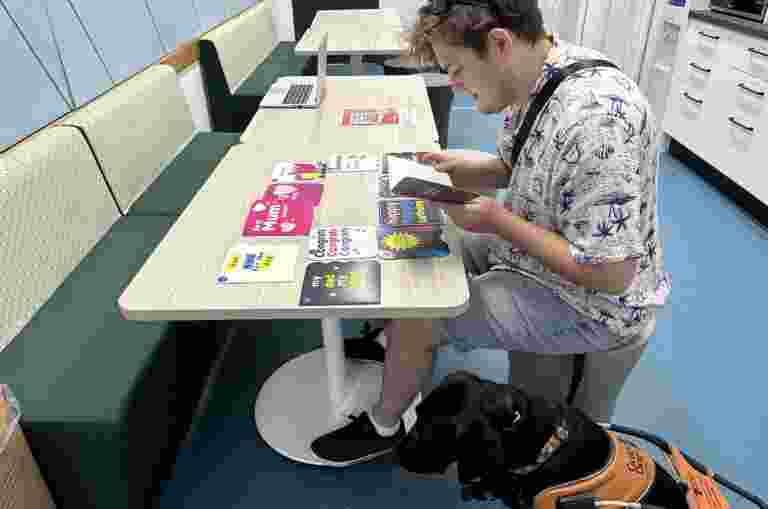 A male with low vision or blindness reads a braille card while sitting down at a table. Their Guide Dog sits at their feet.