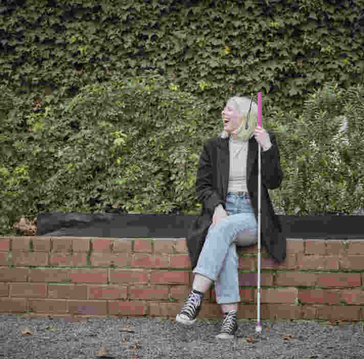 Guide Dogs Client Lucy sits smiling on a brick wall. She holds a white cane.
