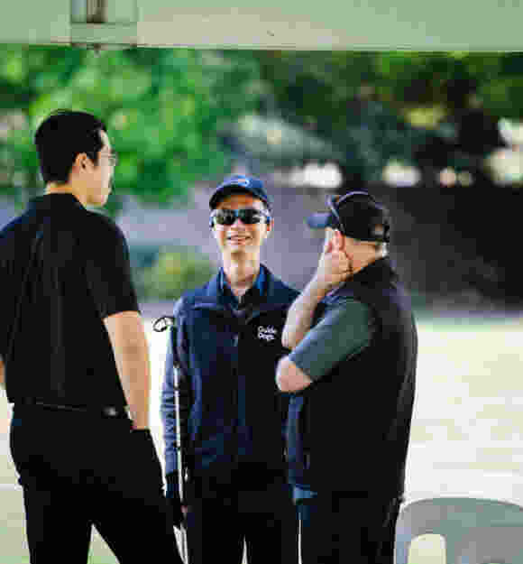 Guide Dogs client Simon stands talking to people outside on his left and right while smiling.