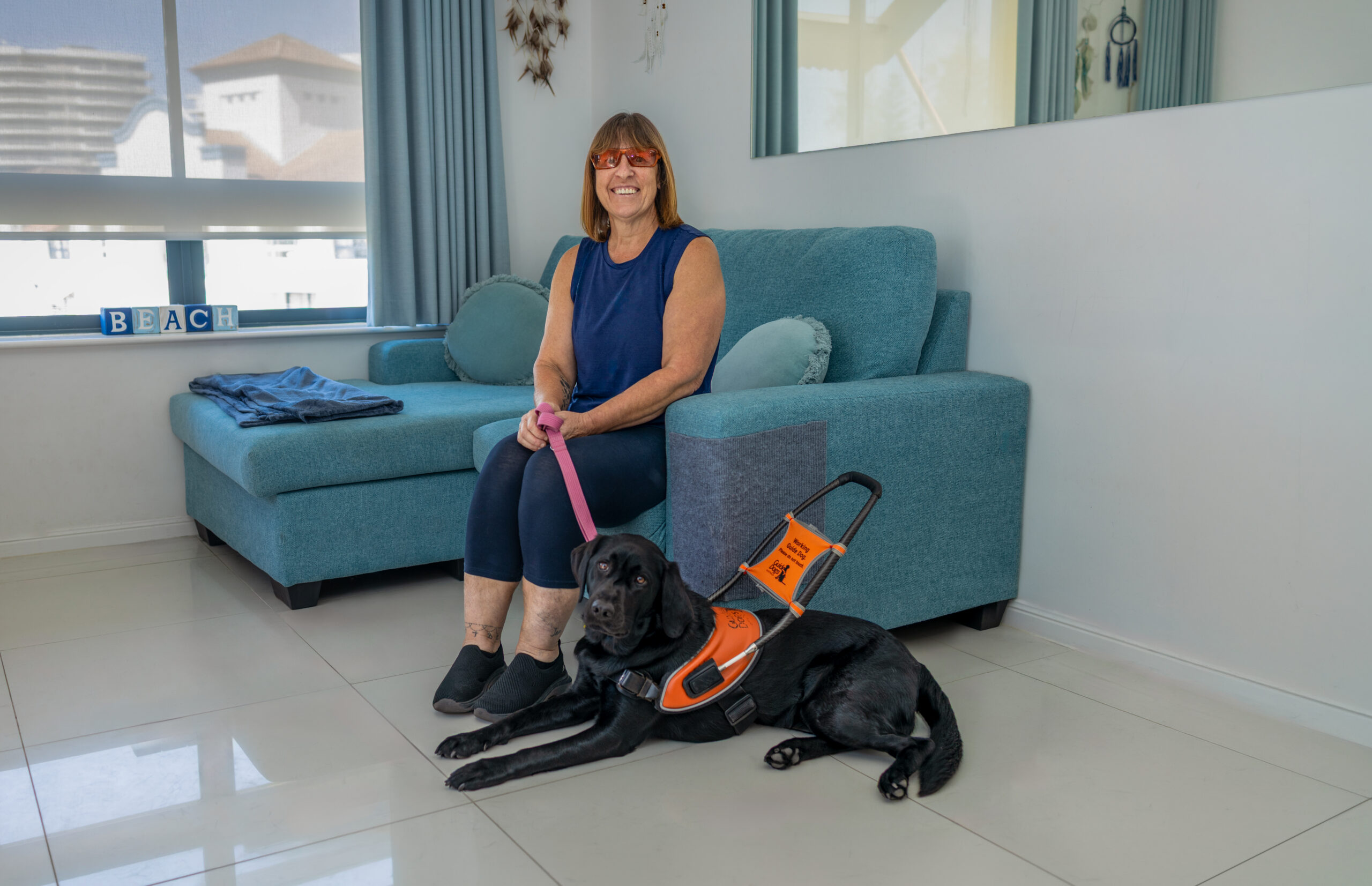 Image shows a woman sitting in an apartment on a blue sofa with a black guide dog in harness at her feed
